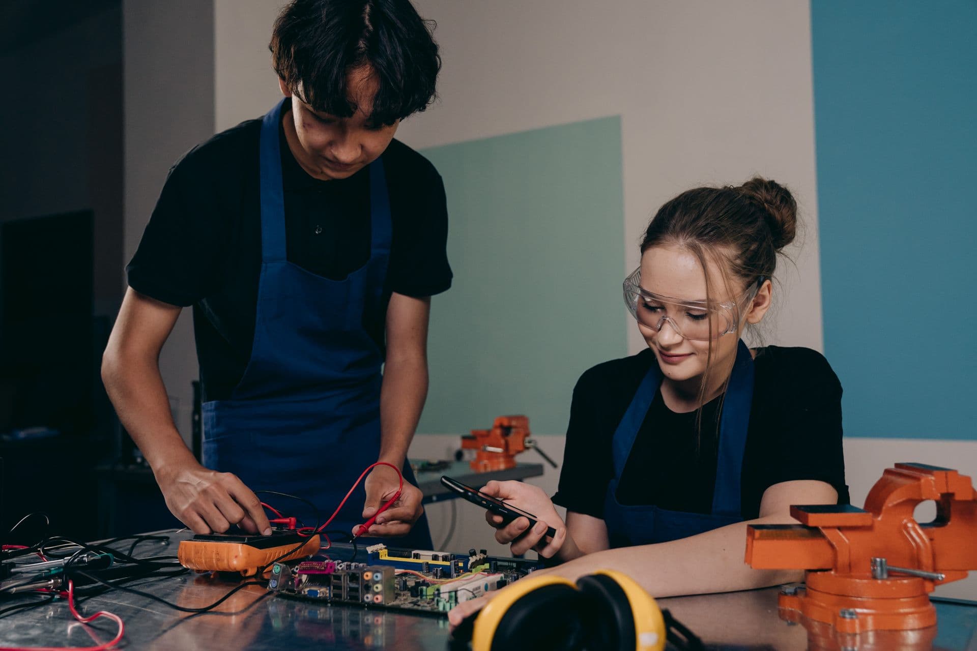 Two young researchers testing a circuit board with a multimeter at a lab workbench.